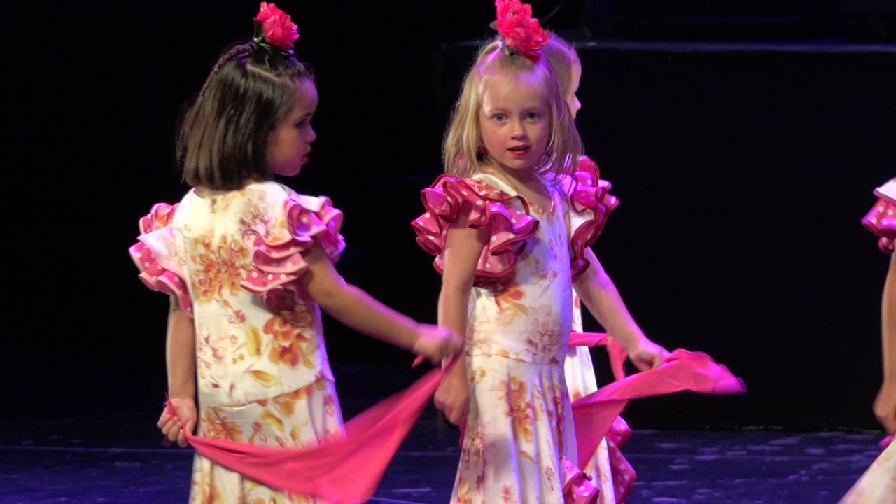 Two young girls in floral dance costumes hold pink fabric on a dark stage.