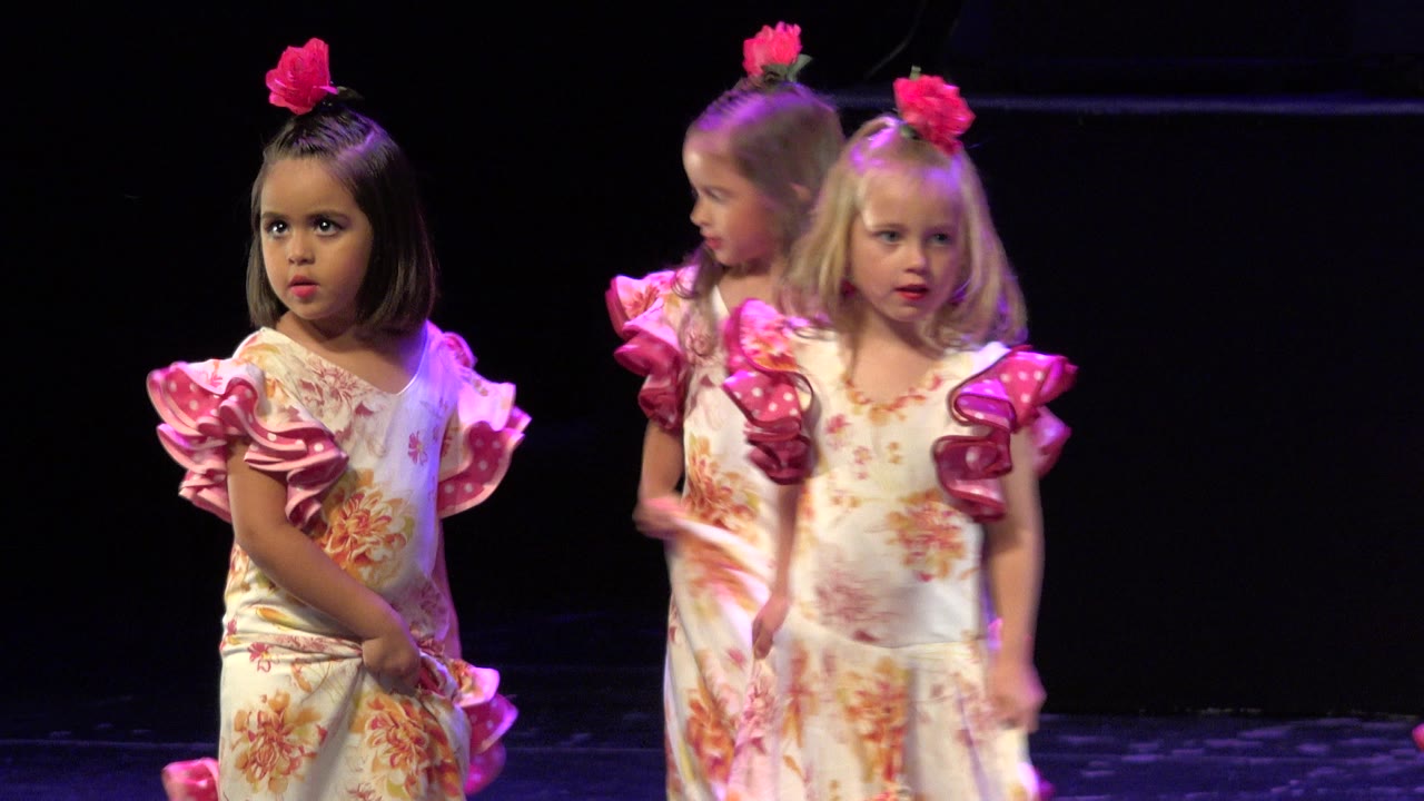 Three young girls in floral dresses with bright pink ruffled sleeves stand on a dark stage during a dance performance in Cabo San Lucas, Mexico.