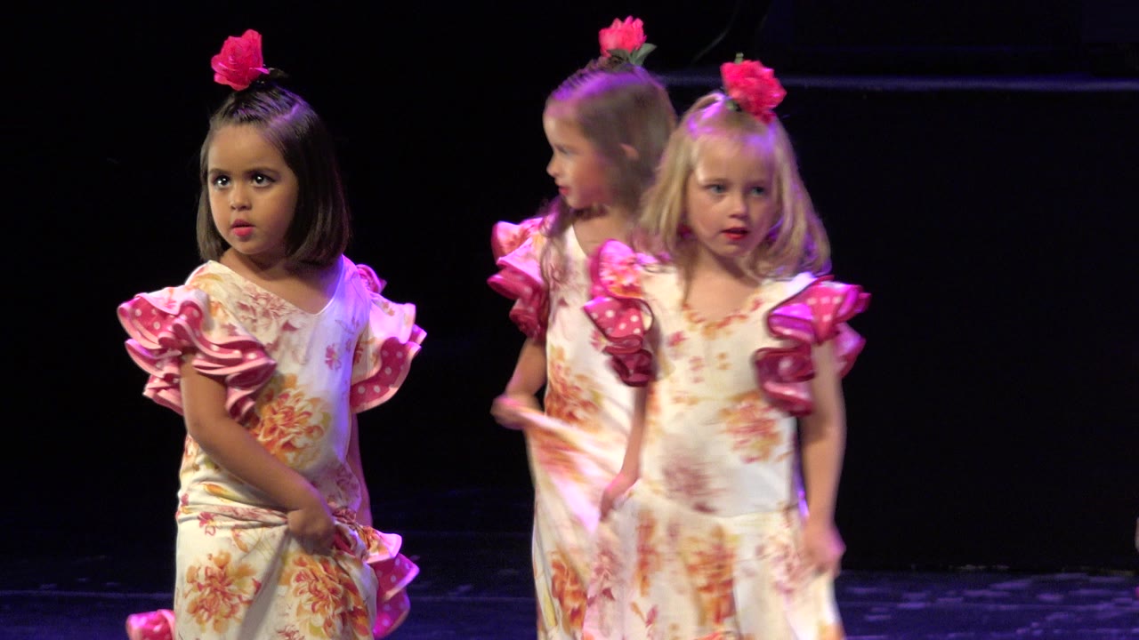 Two girls face forward while another turns sideways in matching floral dresses under purple lighting on a dark stage in Cabo San Lucas, Mexico.