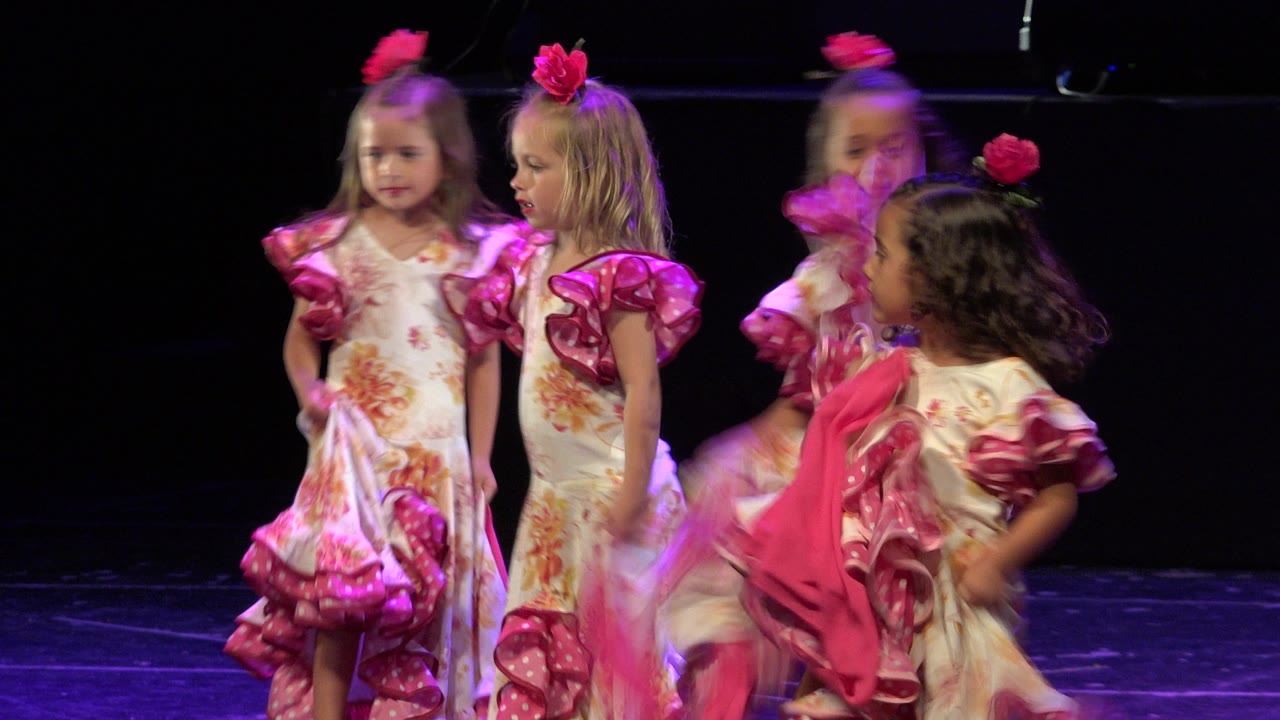 Young girls in ruffled floral dresses dancing on a dark stage.