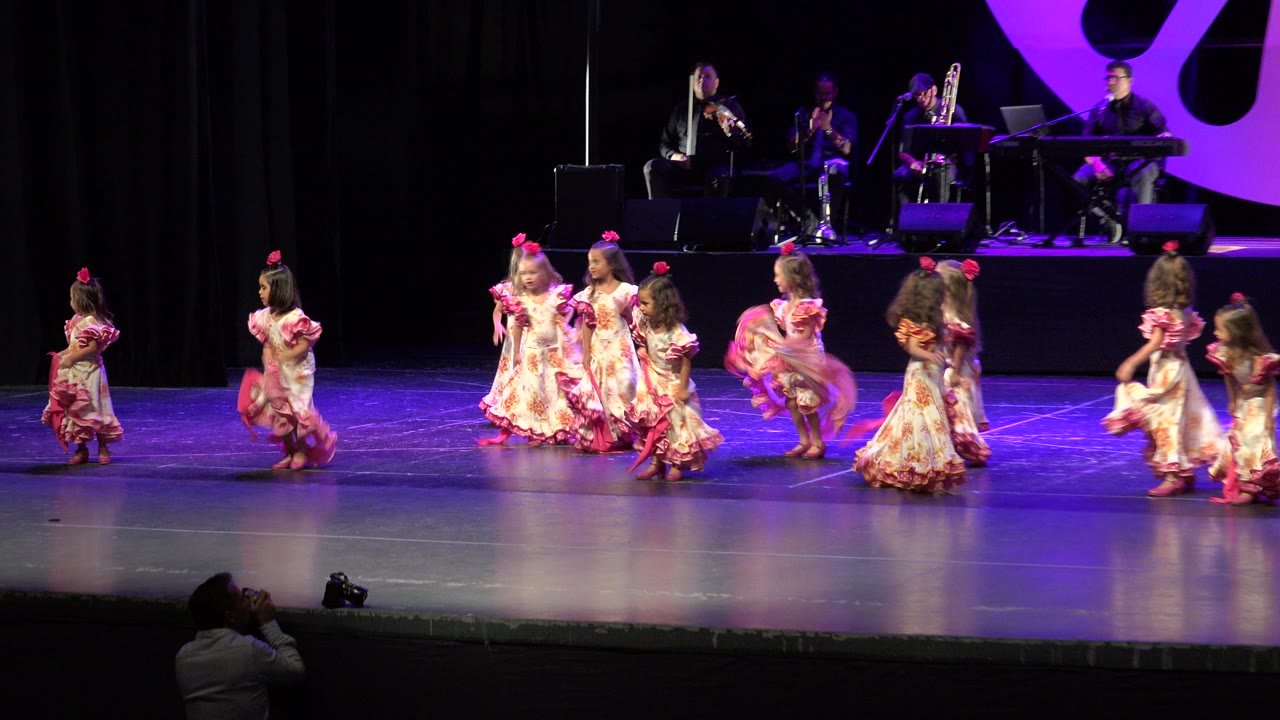 Young girls perform in pink and cream ruffled dresses on a Cabo San Lucas stage while musicians sit on a raised platform under purple lighting.