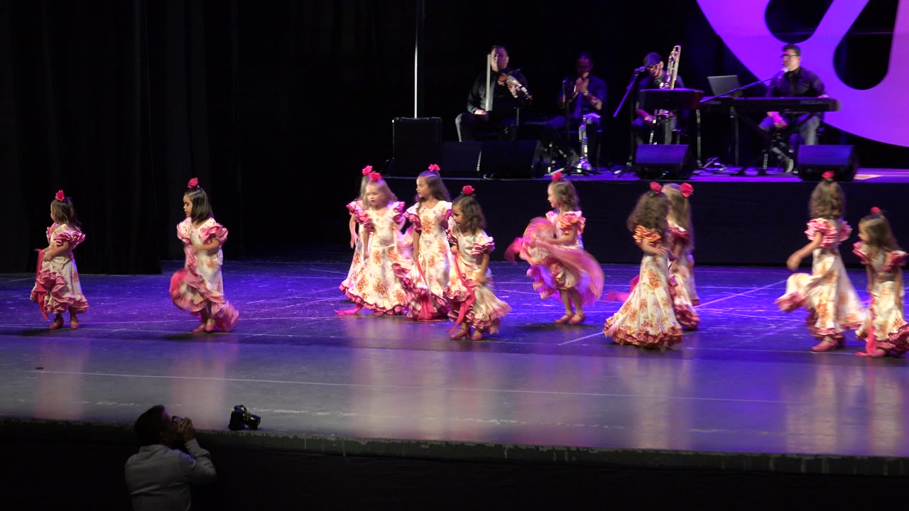 The group of girls continues the Cabo San Lucas stage routine, clustered near center while other dancers enter from the sides and the band remains in the background.