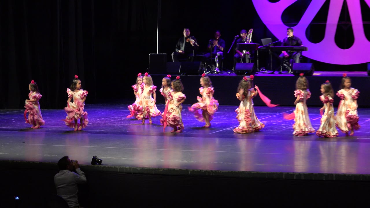 Girls in floral dresses dance in a line across the stage under purple lighting.