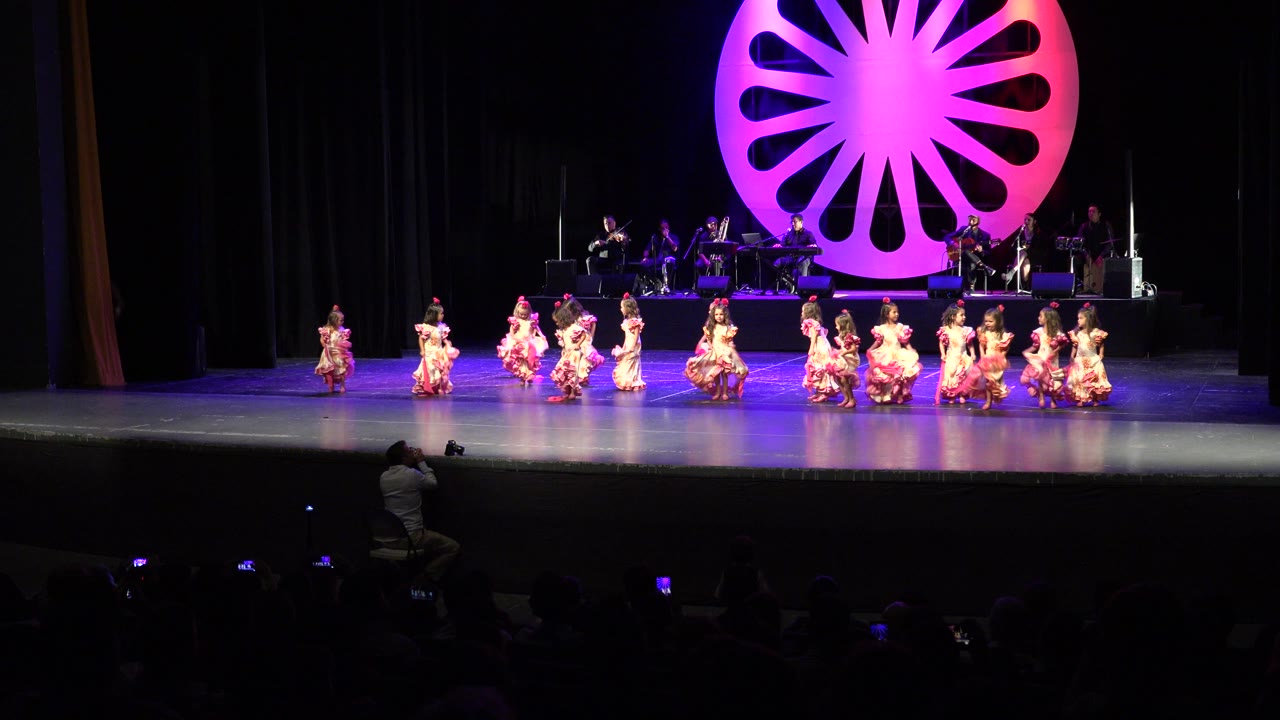 Wide audience view in Cabo San Lucas, Mexico, showing children in pink ruffled dresses performing across a stage while musicians sit behind a large pink circular backdrop.