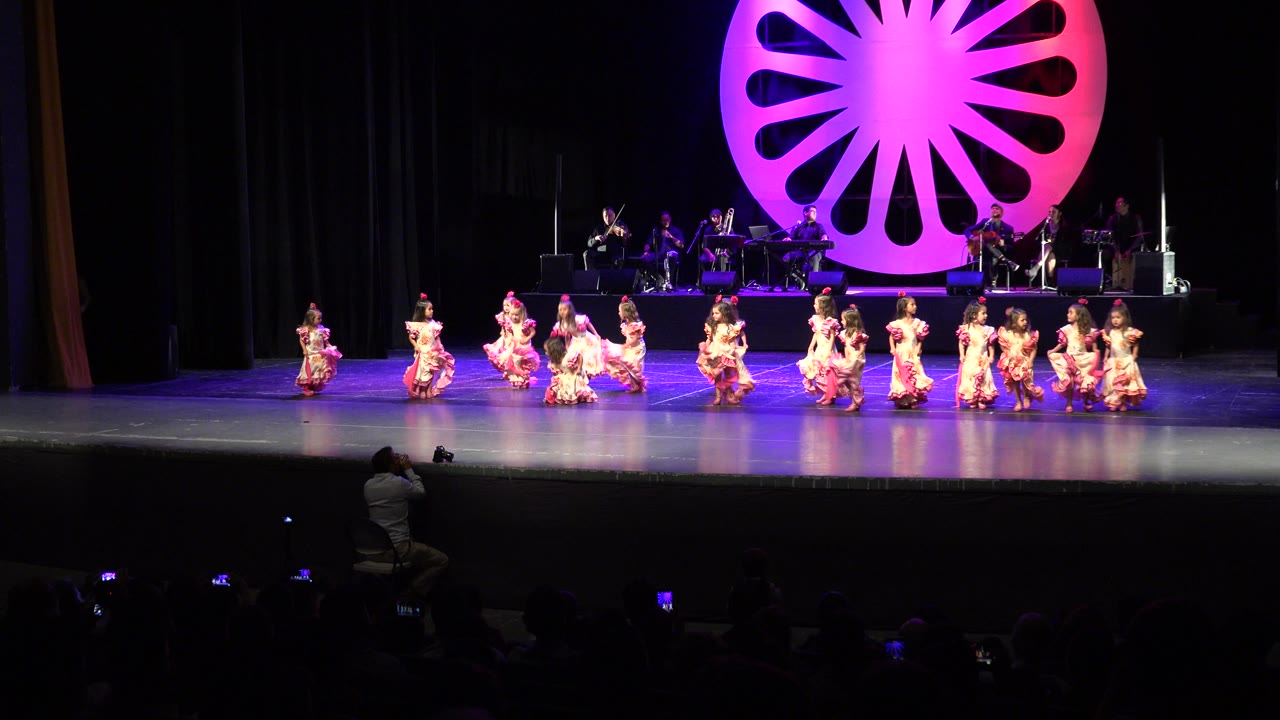 A line of children in pink dresses moves across the stage under purple light.