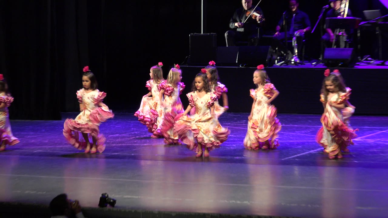 A wide Cabo San Lucas performance frame shows several young girls moving across the stage in matching pink-trimmed dresses while seated musicians remain visible above them.