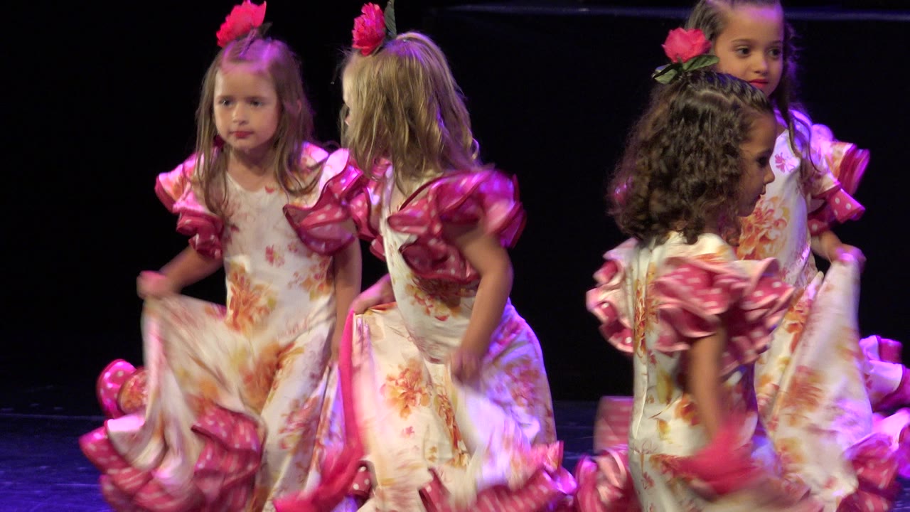Little girls in white floral dresses with pink ruffled sleeves stand and move together on a dark stage in Cabo San Lucas, Mexico.