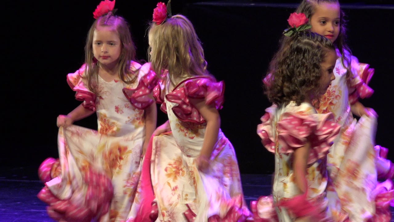 Several young girls in matching floral dresses turn and pass one another under purple stage light during the Cabo San Lucas performance.