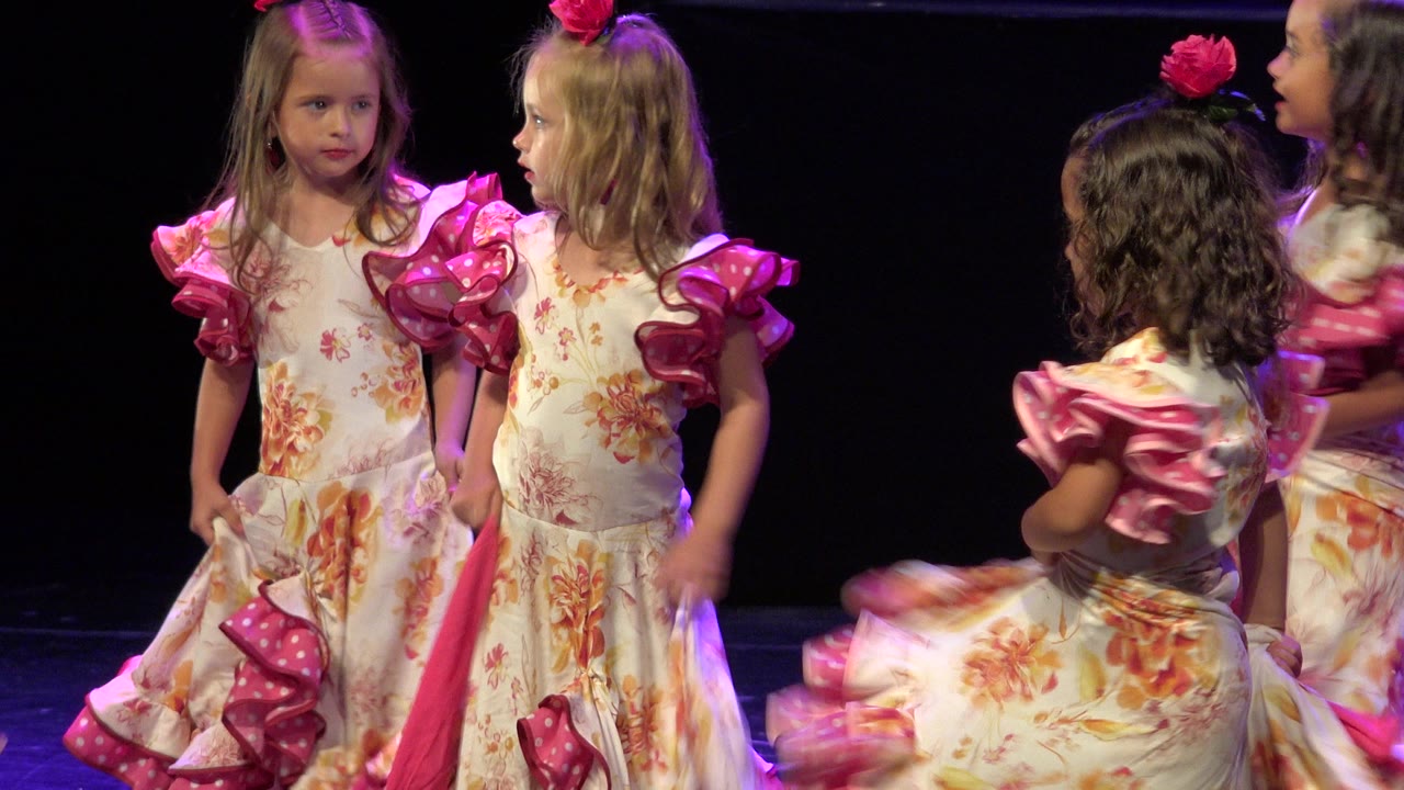 Young girls in floral dresses with pink ruffled sleeves stand close together on a dark stage in Cabo San Lucas, Mexico, while another child moves through the foreground.