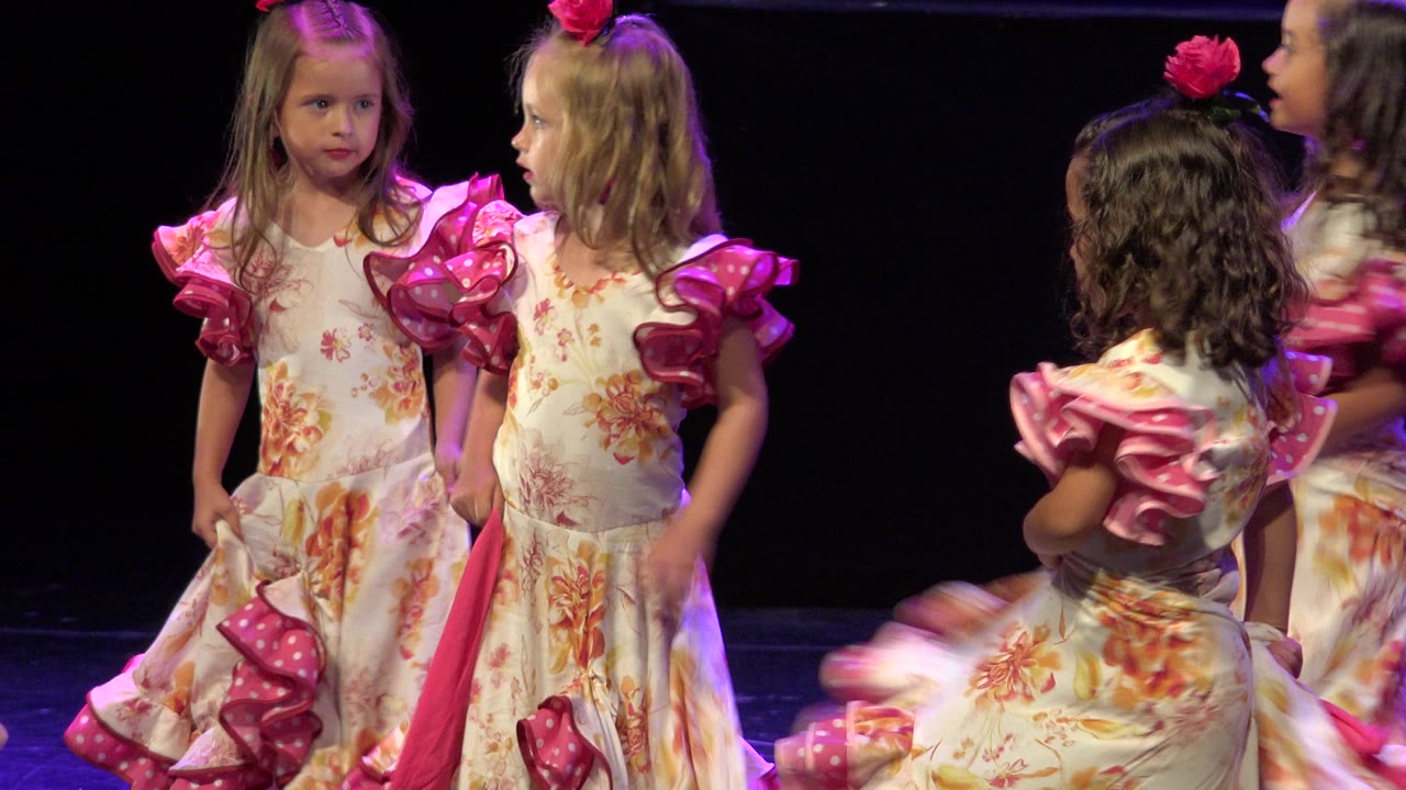 Two girls in floral folk dance dresses face each other at center stage in Cabo San Lucas, Mexico, with a child in a similar dress blurred in the foreground.