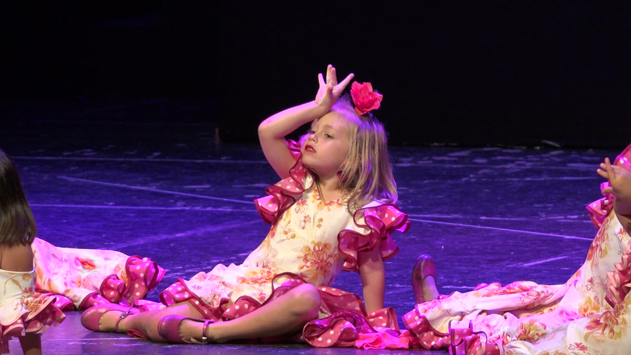 At 0:00, a young girl in Cabo San Lucas sits on the stage floor in a floral dress with pink ruffles, posing with one hand near her forehead while another child is partly visible beside her.