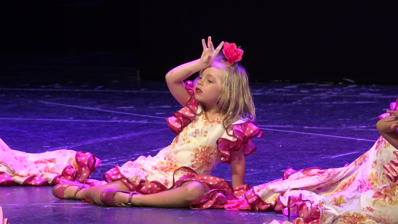 Small girl in a pink stage dress sits with one hand raised near her face.