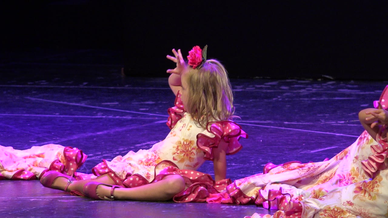 In Cabo San Lucas, the seated girl remains on the stage floor with one hand raised near her face, pink ruffled sleeves and skirt trim catching the purple stage light.