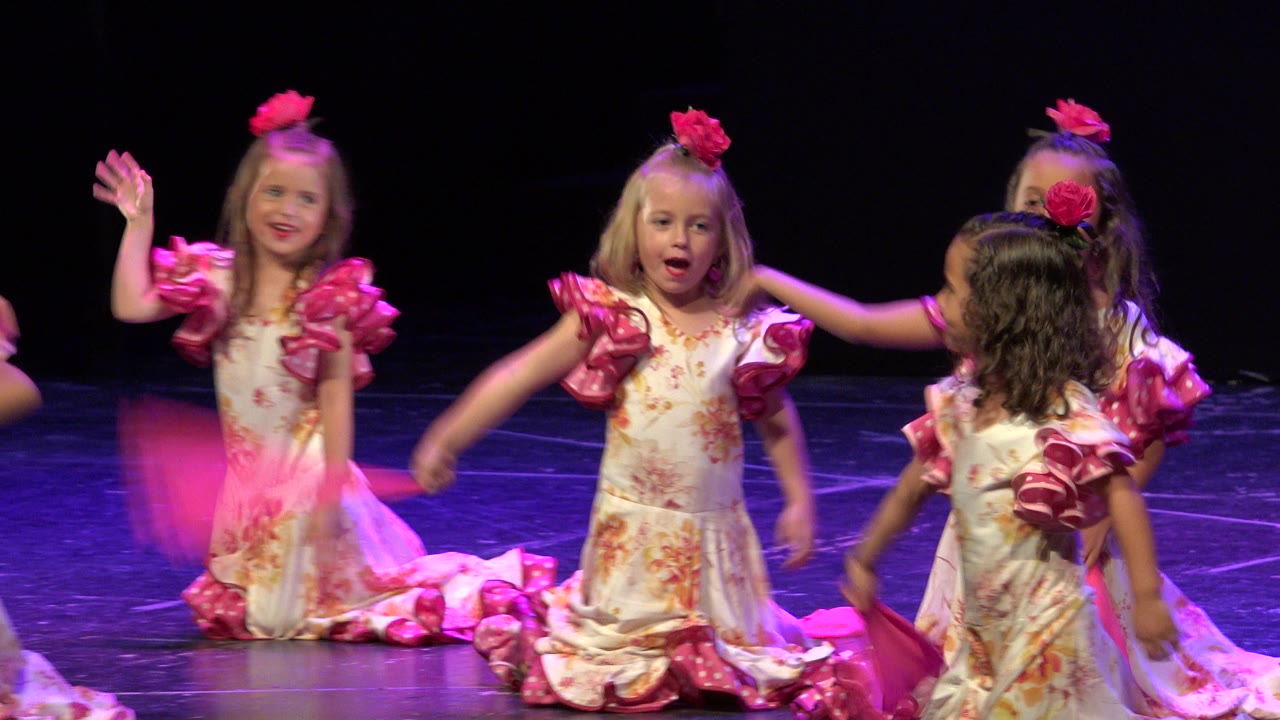 Little girls in pink-trimmed dresses dancing on a dark stage.