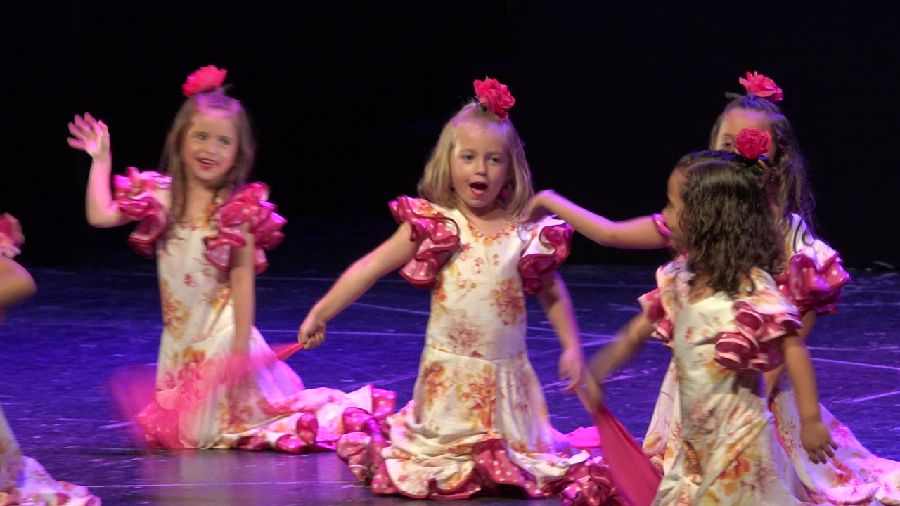 A blonde girl in a pink ruffled dress waves near center frame while other children in matching costumes move around her on the Cabo San Lucas stage.