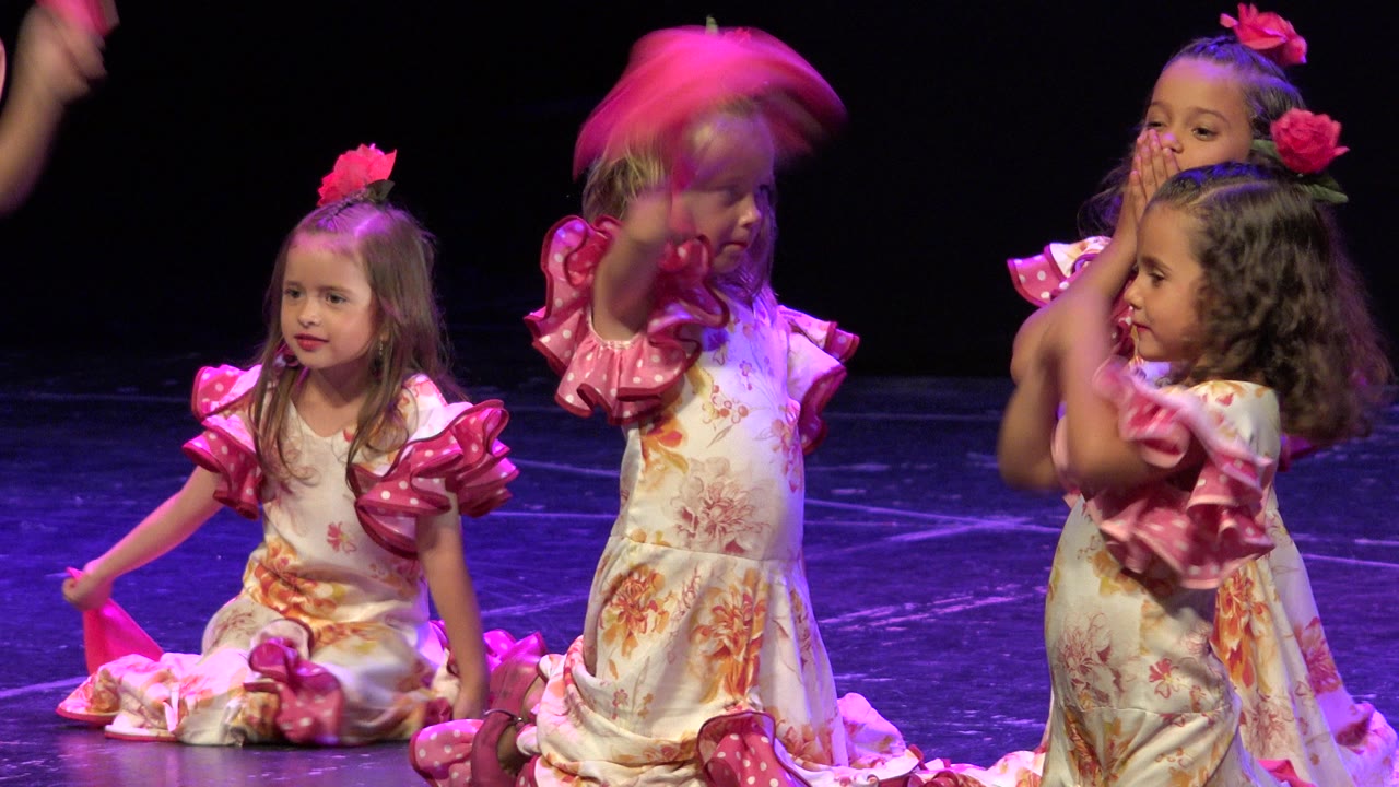 Children in floral dresses with shiny pink sleeves handle bright pink scarves on a dark performance stage in Cabo San Lucas, Mexico.