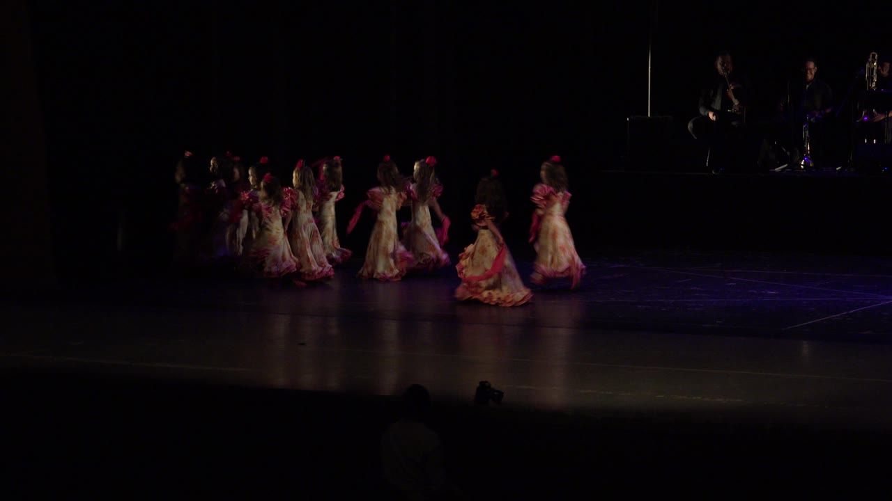 The group of children in red and white costumes moves across the dark performance stage in Cabo San Lucas, Mexico, while musicians remain dimly lit in the background.