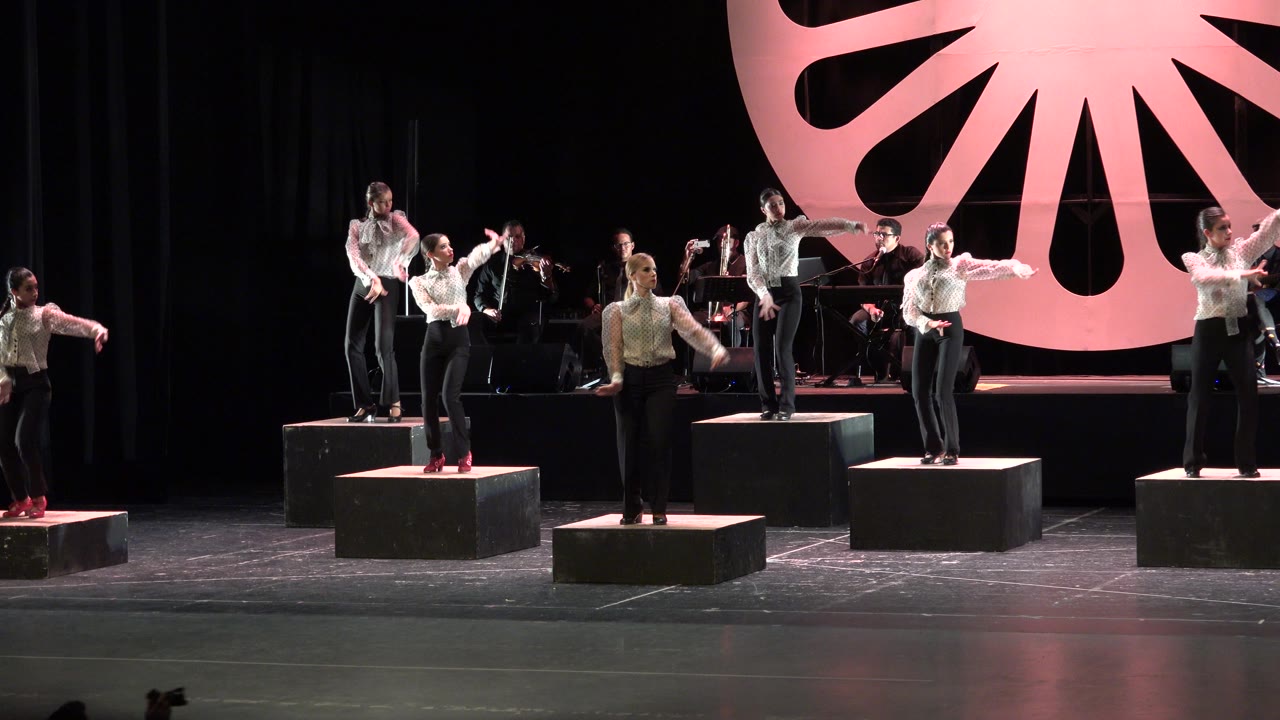 The Cabo San Lucas stage performance continues with several performers extending their arms from separate risers as the seated band remains visible behind them.