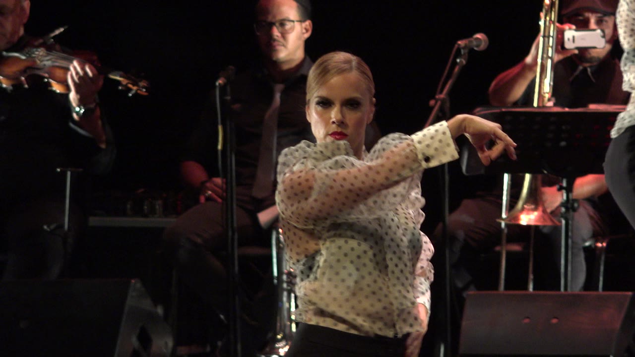 Female dancer points across a dark stage with musicians behind her.