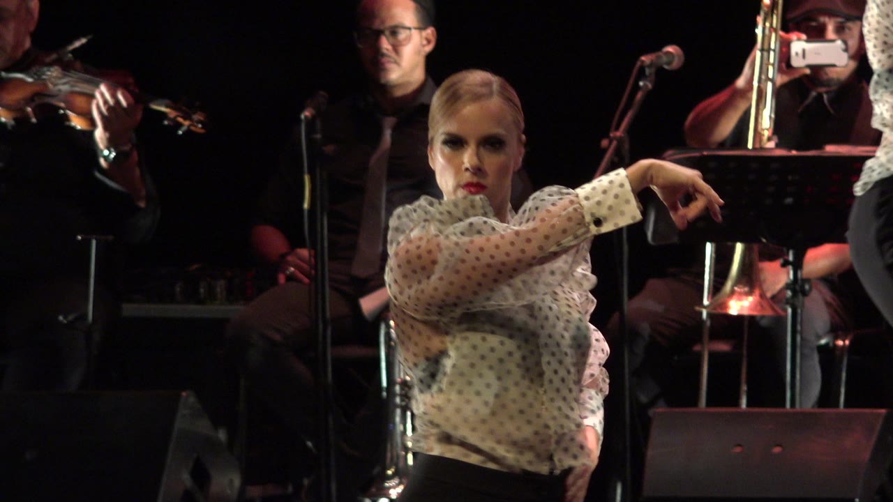 In Cabo San Lucas, Mexico, the dancer maintains a strong forward-facing pose with one arm extended as musicians and stage microphones remain visible behind her.