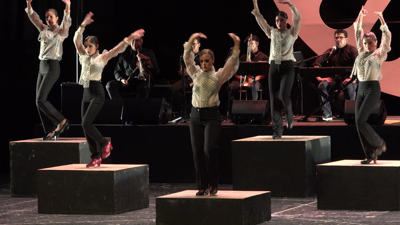Five women dance on stage blocks with musicians behind them.