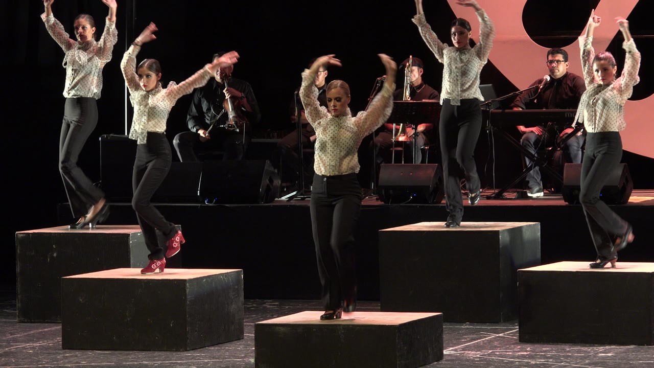 The dancers hold high arm positions on separate black platforms in Cabo San Lucas, Mexico, while the band remains seated in the dark background.