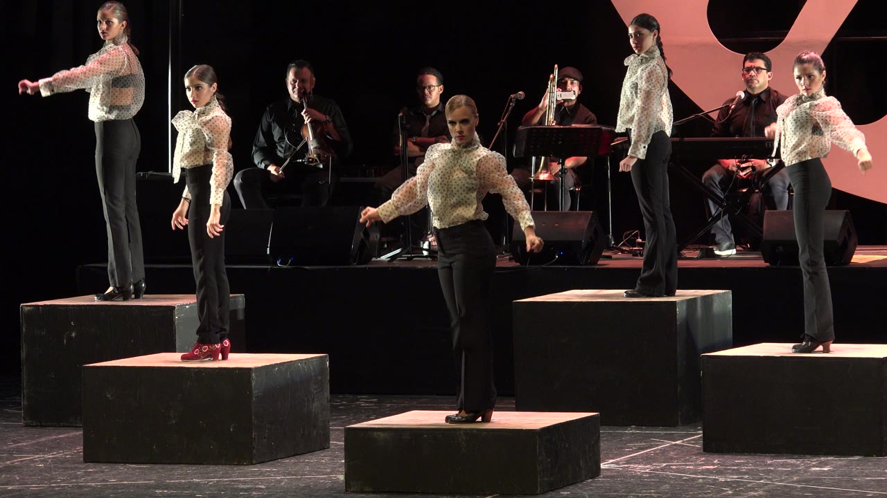 The group holds a similar formation at 0:01, with one dancer centered in front and other performers standing on risers in the Cabo San Lucas stage setting.