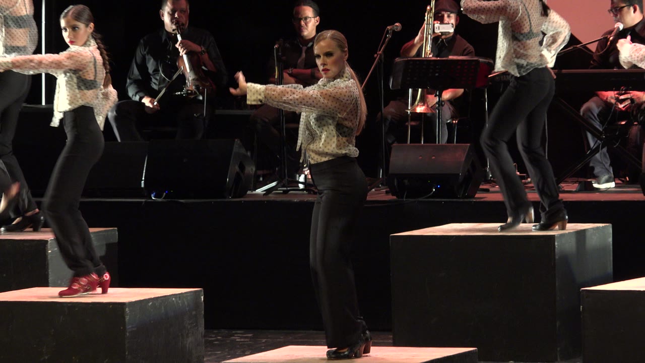Blonde dancer extends an arm on a dark stage with musicians behind her.