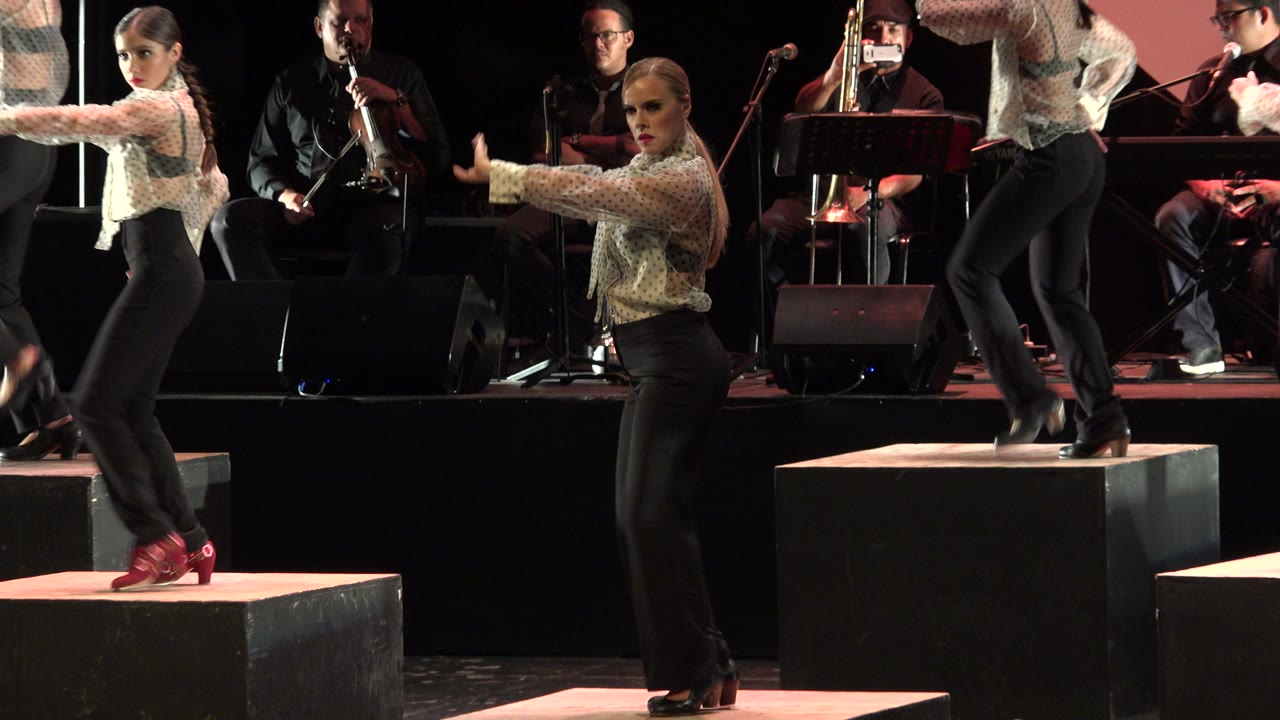 In Cabo San Lucas, Mexico, the dancer faces the camera with one arm extended as another performer steps across a raised black platform near the band.