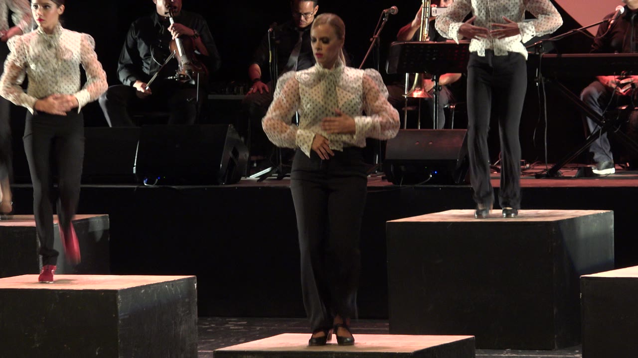 Women tap dancing on black stage platforms with musicians behind them.