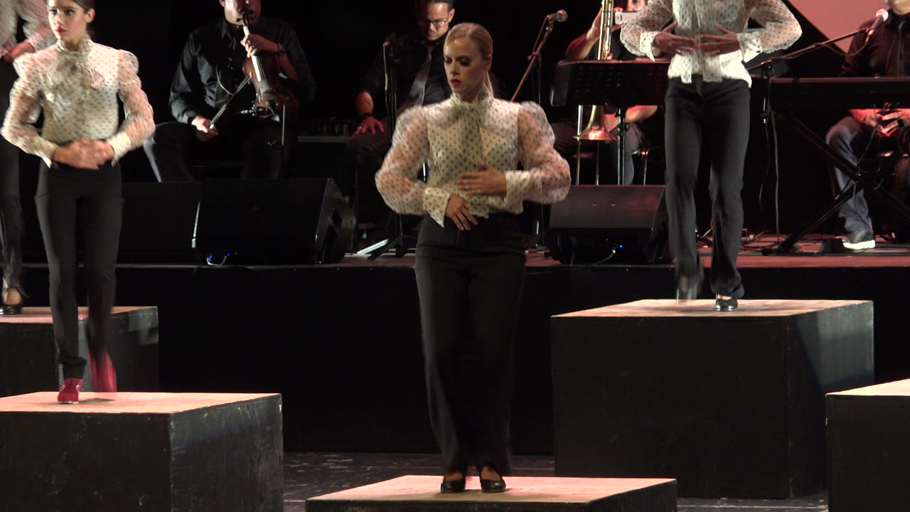 A woman in red tap shoes steps on a front platform in Cabo San Lucas, Mexico, while another dancer stands on a rear riser near the band.