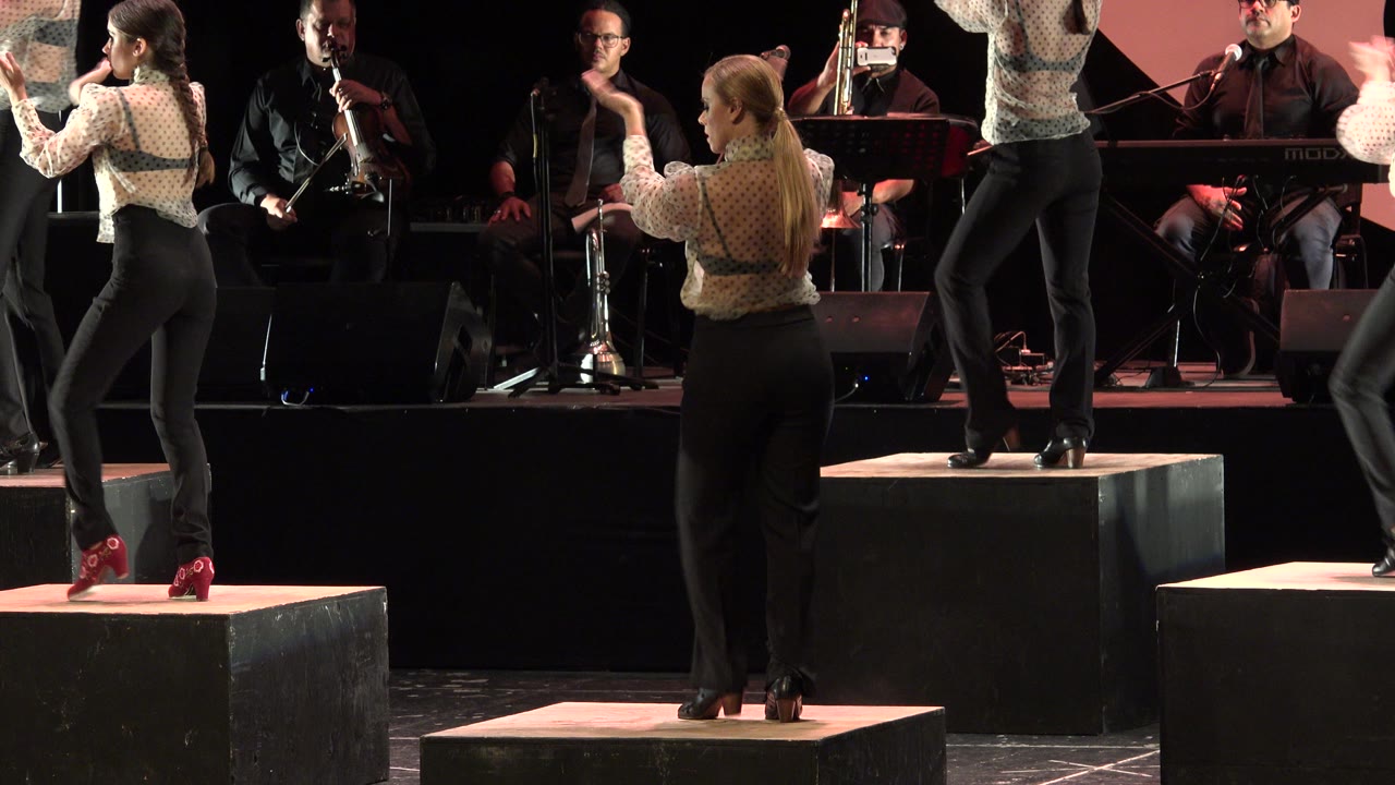 One second in, two women face each other while clapping on separate stage blocks, with seated musicians and instruments visible behind them in Cabo San Lucas.