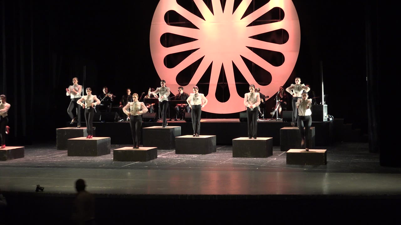 Wide stage with male dancers clapping on platforms before a red circular backdrop.