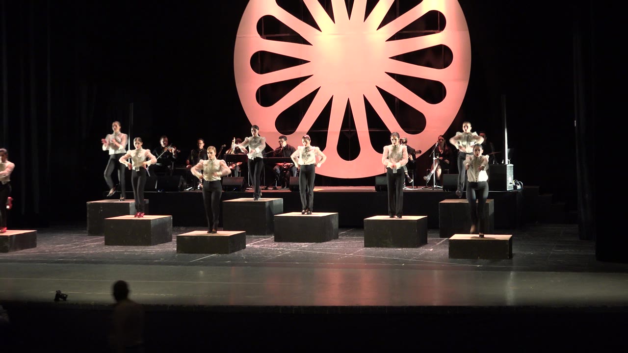 Cabo San Lucas performers continue clapping in formation on separate black platforms while the seated band remains visible behind the front line.