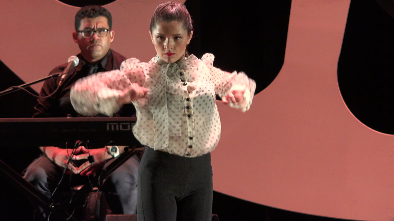 In Cabo San Lucas, Mexico, a young dancer in a white patterned blouse faces forward while a seated keyboard player and microphone sit behind her on the pink and black stage.