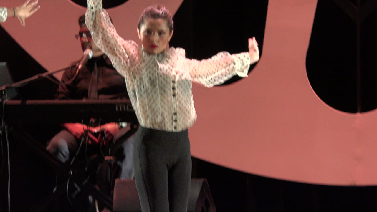In Cabo San Lucas, Mexico, the dancer remains centered with one arm lifted and one arm extended, with a keyboard player and stage equipment visible in the background.