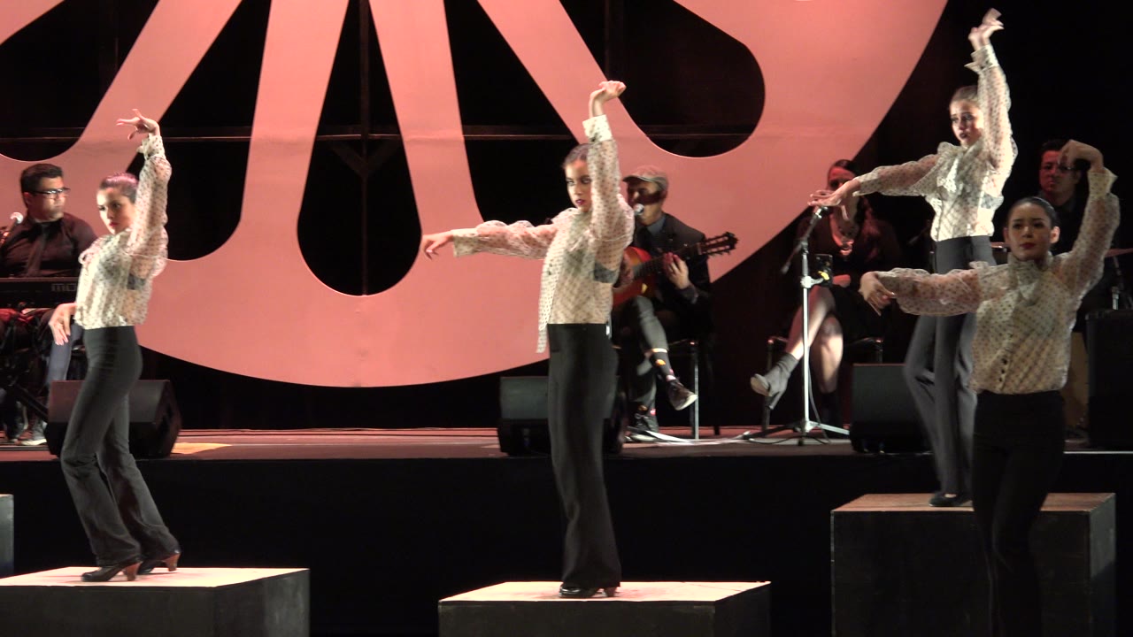 Young female dancers begin a choreographed stage pose in Cabo San Lucas, Mexico, with seated musicians and large pink backdrop shapes behind them.