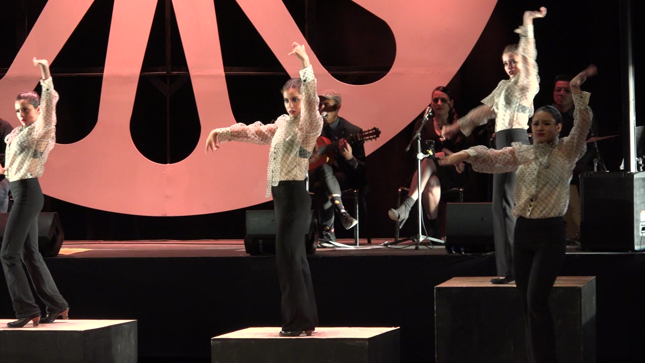 Three dancers perform synchronized gestures in front of a pink stage backdrop.