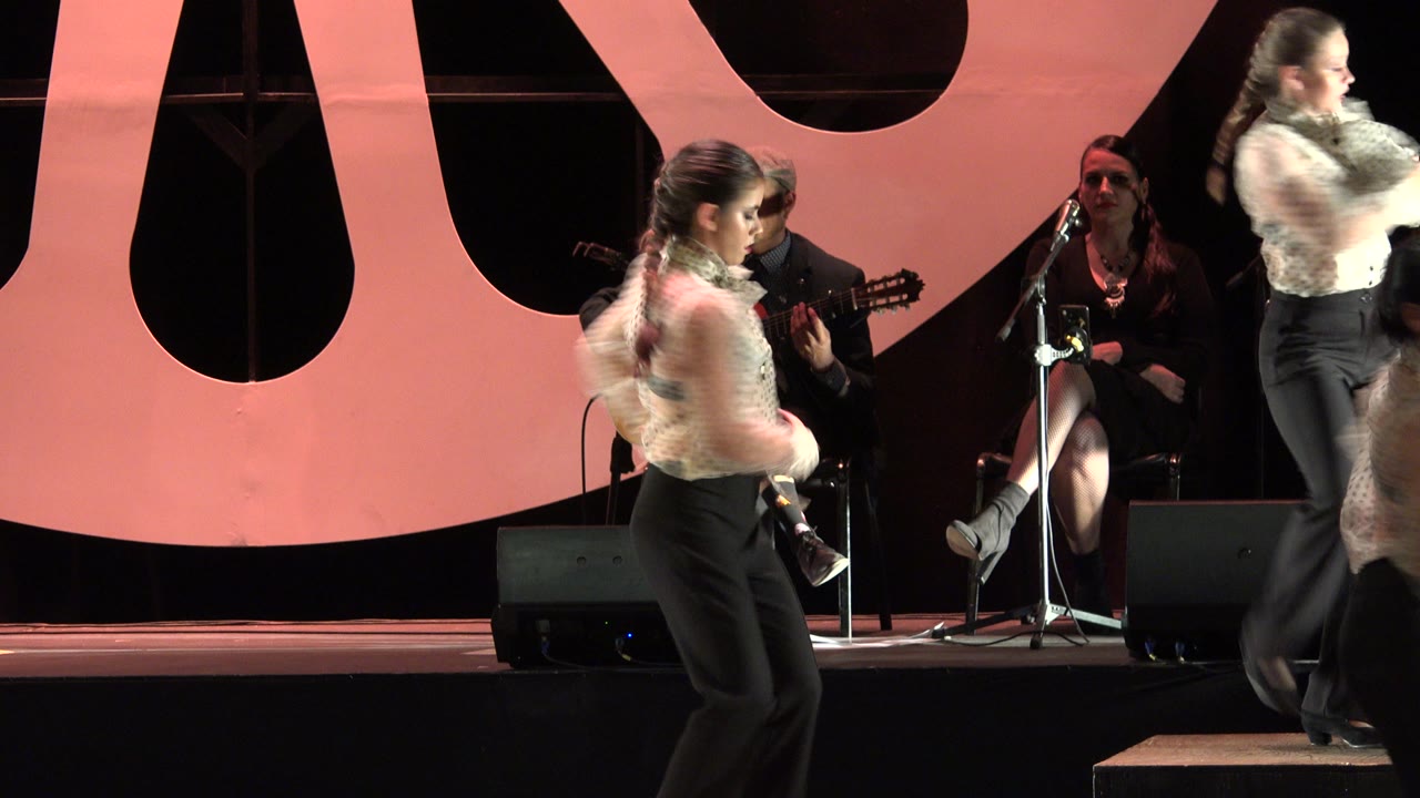In Cabo San Lucas, Mexico, one dancer turns near center frame as another passes at right, with guitar players, a singer, microphones, and floor speakers in the background.