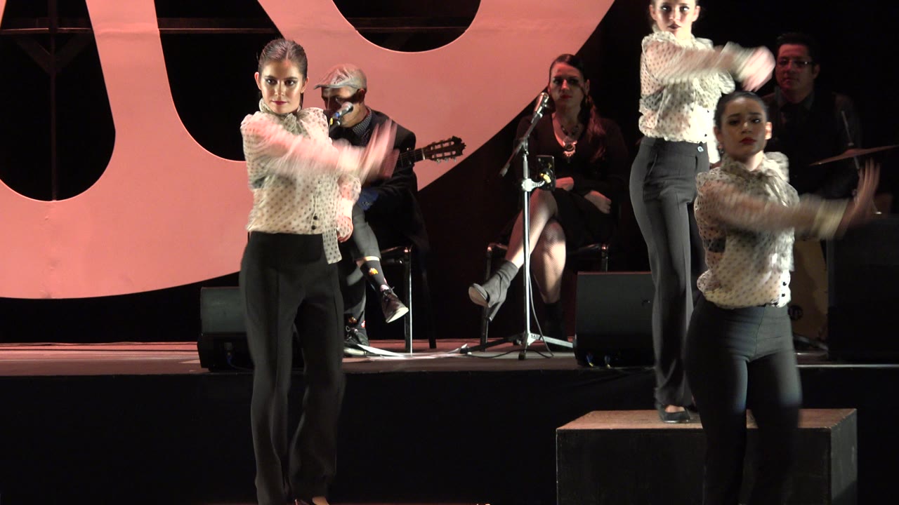 Women tap dancers in suits perform on a stage with musicians behind them.