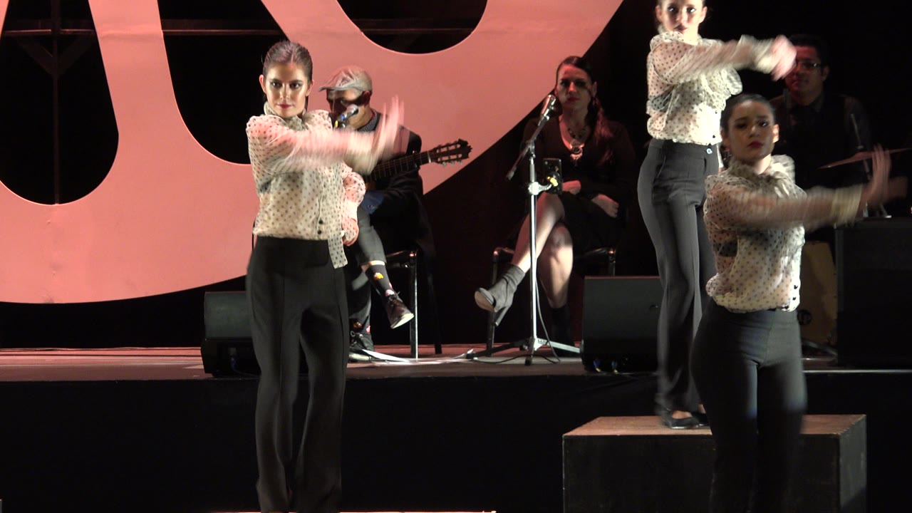 The Cabo San Lucas stage performance continues with women in suits extending their arms while musicians sit in the dim background near microphone stands.