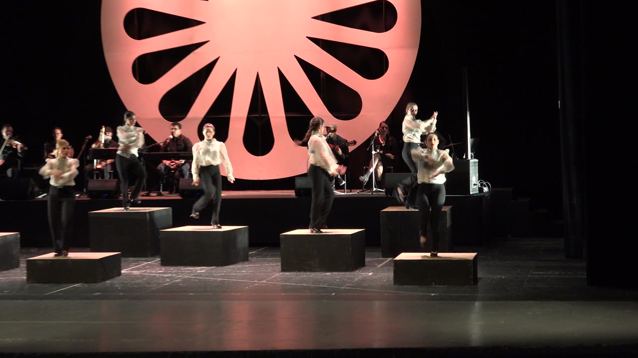 Dancers on stage platforms in front of musicians and a large lit wheel backdrop.