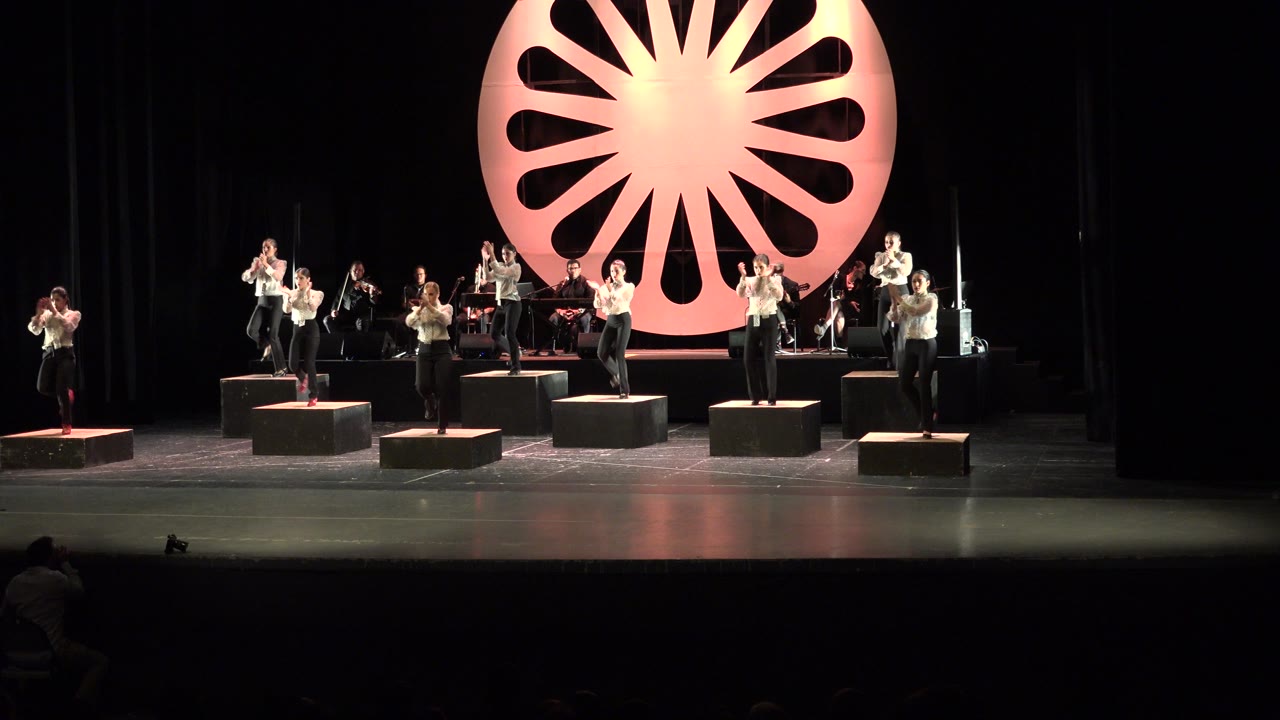 In Cabo San Lucas, Mexico, nine adult dancers stand across black risers on a dim stage with musicians behind them and a glowing orange circular prop centered in the background.