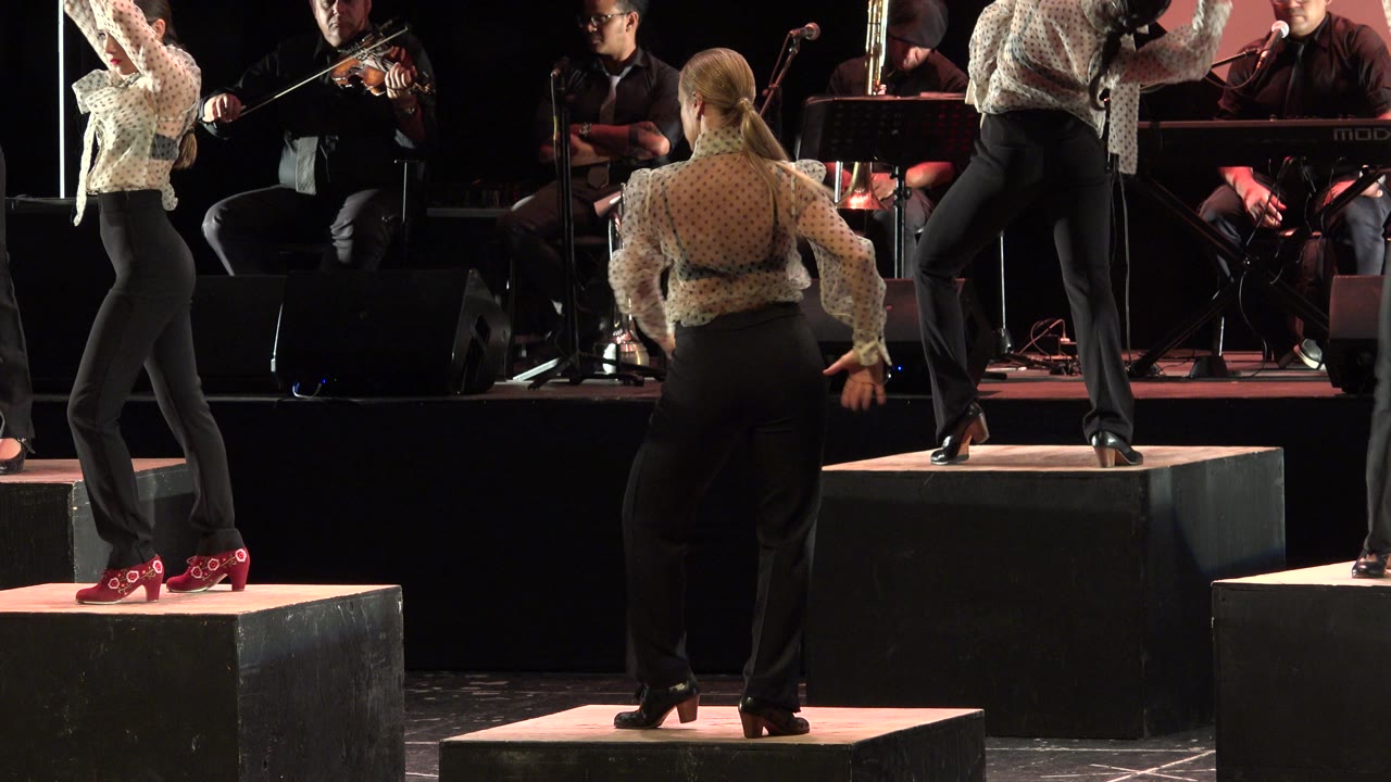 In Cabo San Lucas, Mexico, female dancers stand on black stage risers while seated musicians and instruments fill the dark background.