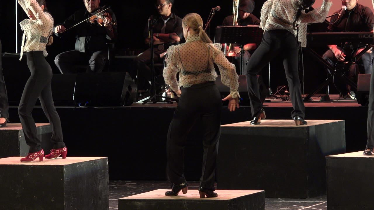 A dancer seen from behind holds her arms low on a raised platform as musicians play across the rear of the Cabo San Lucas stage.