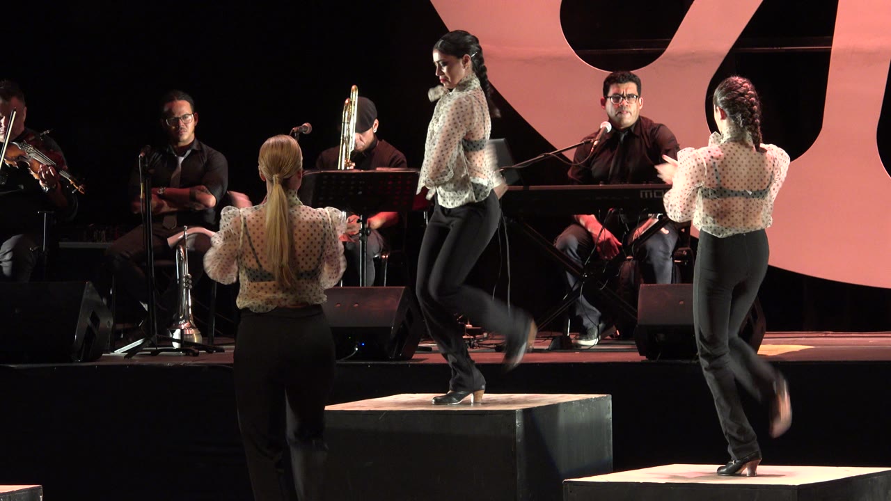 In Cabo San Lucas, Mexico, women in patterned blouses and black trousers perform on lit stage blocks while musicians sit behind them near microphones.
