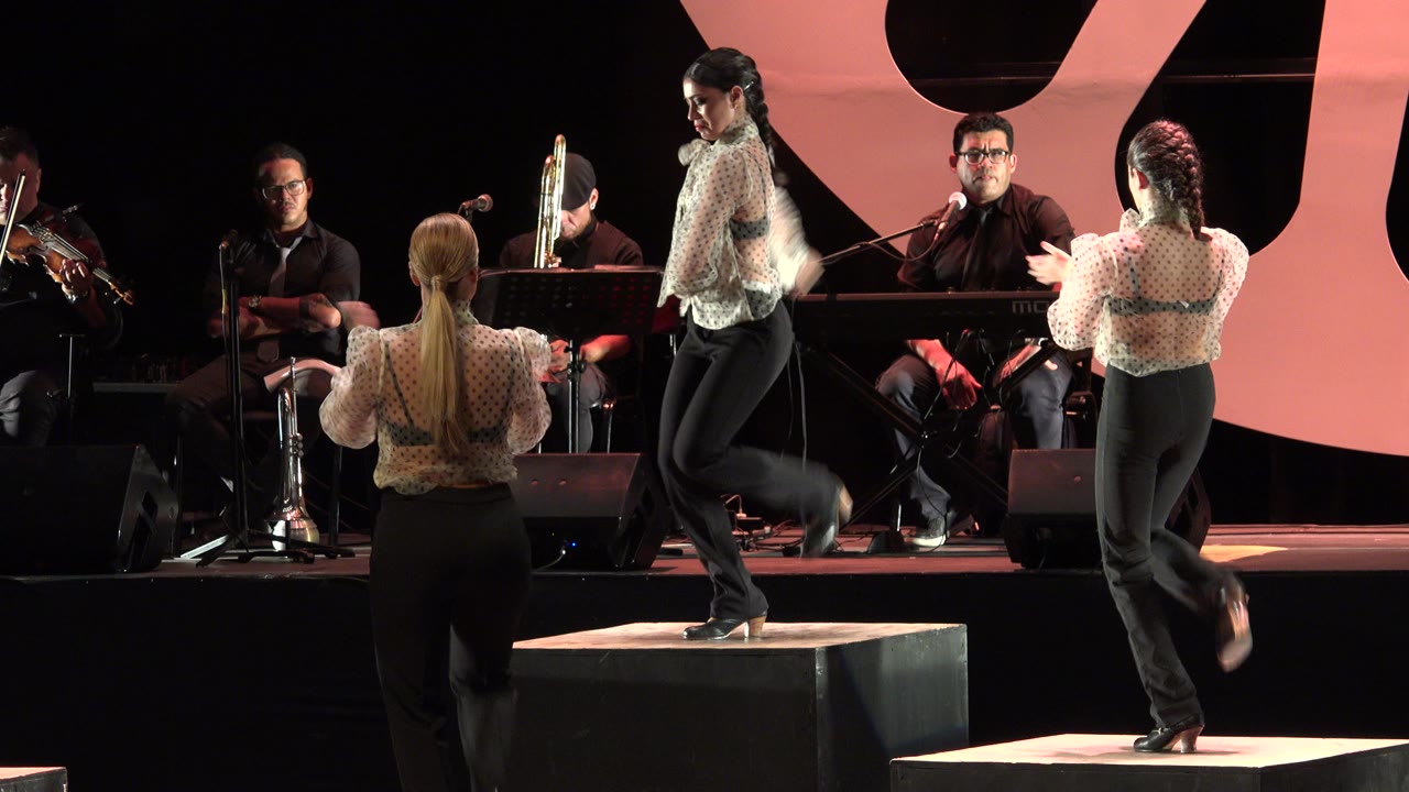 In Cabo San Lucas, Mexico, two women clap near the front while another dancer lifts a leg on a raised platform and the band remains seated in the background.