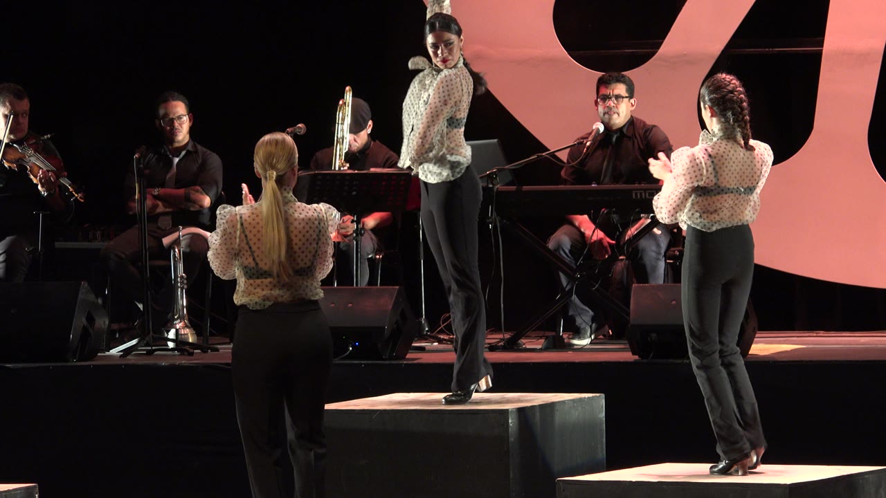Three women perform in formation on a dark stage with a pink backdrop.