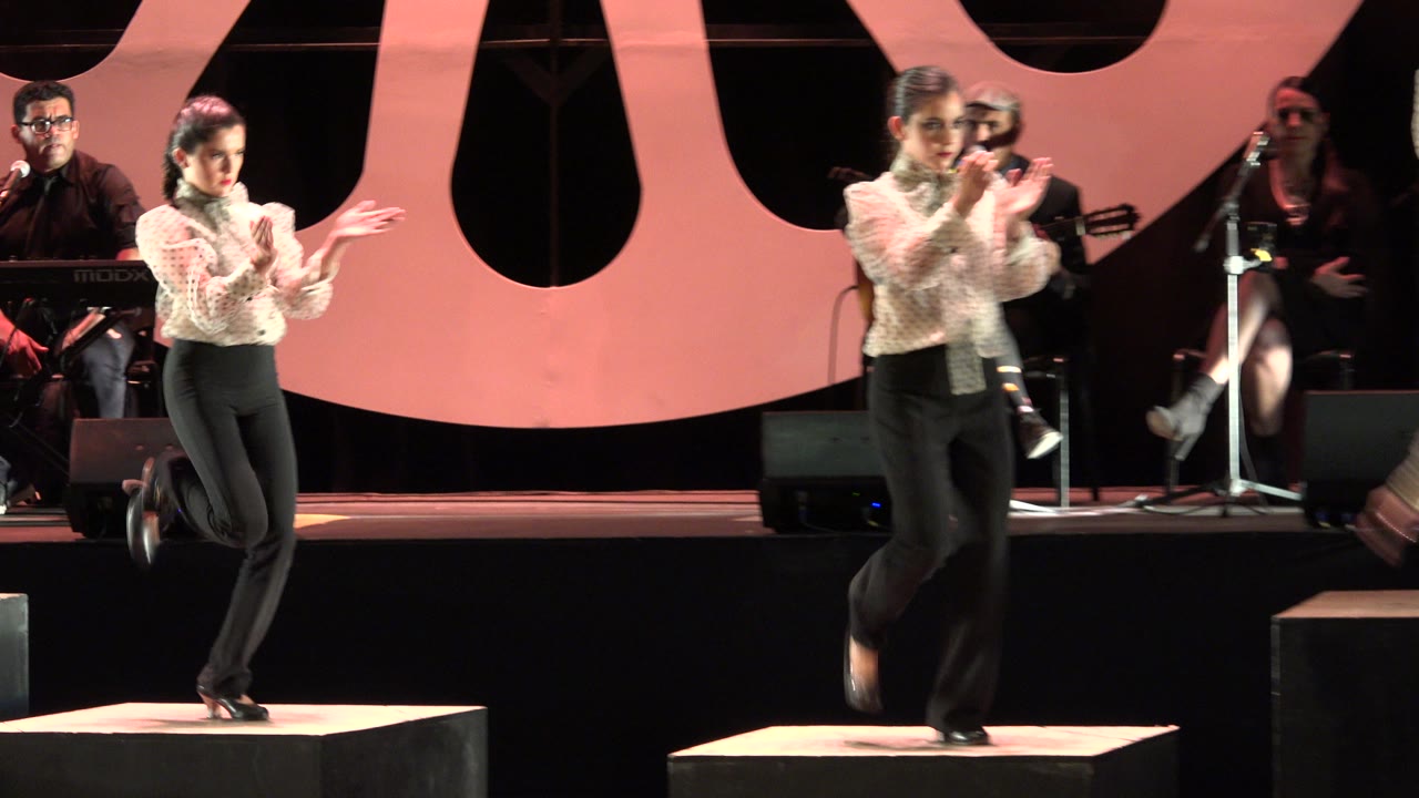 A woman claps on a black platform while another dancer crosses in front of the stage, with musicians and microphone stands visible in the Cabo San Lucas performance setting.