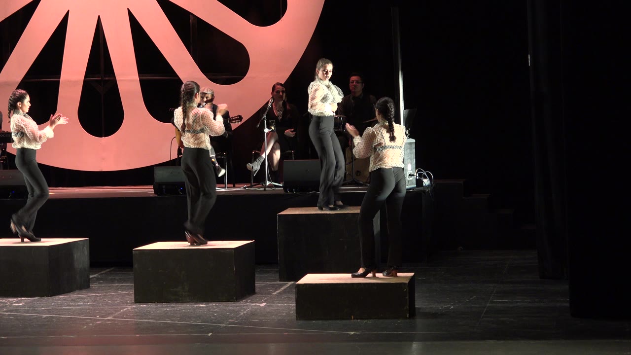 Dancers clap around a soloist on a lit stage with musicians in back.